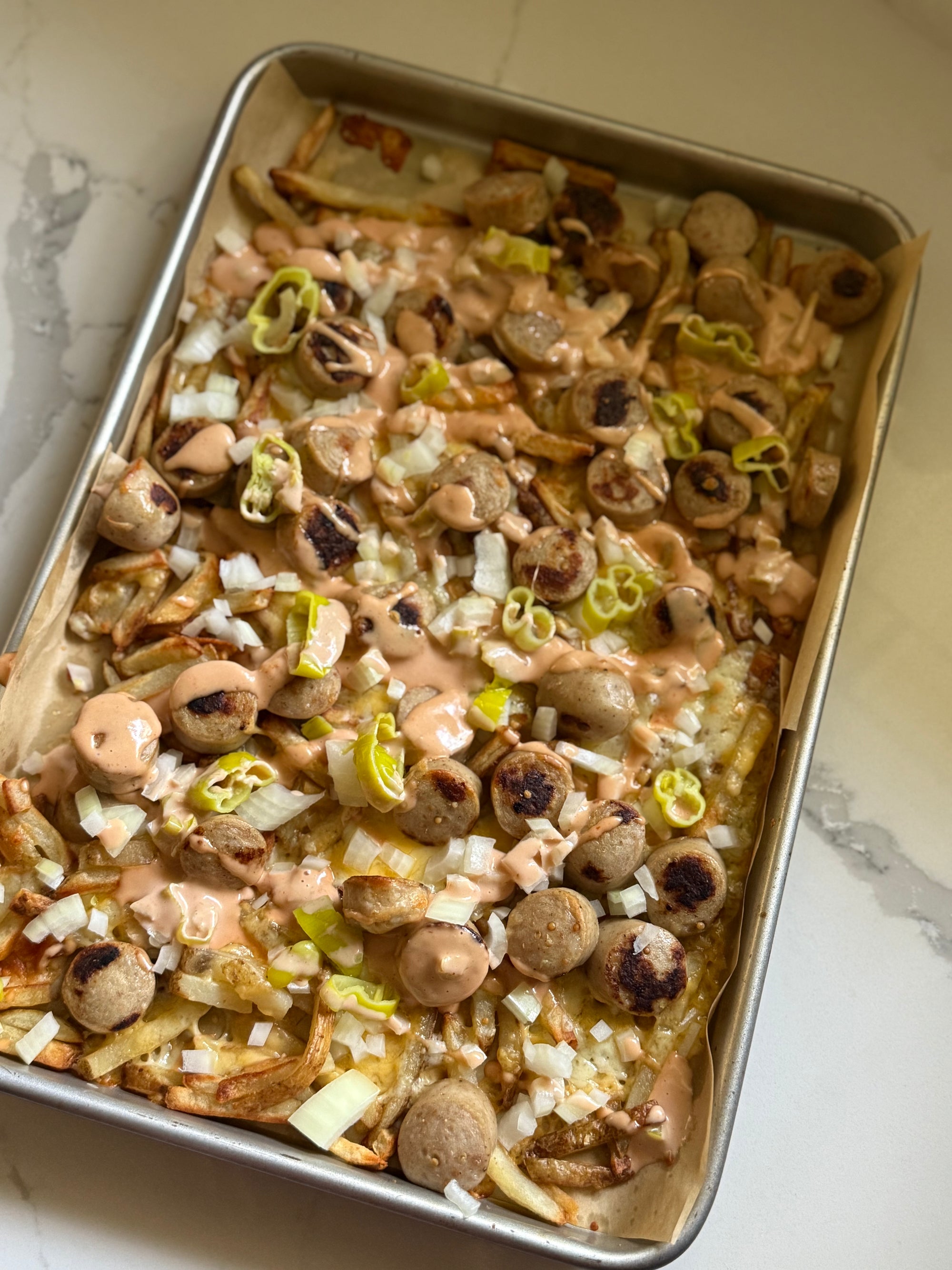 Loaded  fries in a metal baking pan, topped with sliced bratwurst, diced white onions, green onions, and a light pink creamy sauce, photographed from above on a light marble surface.