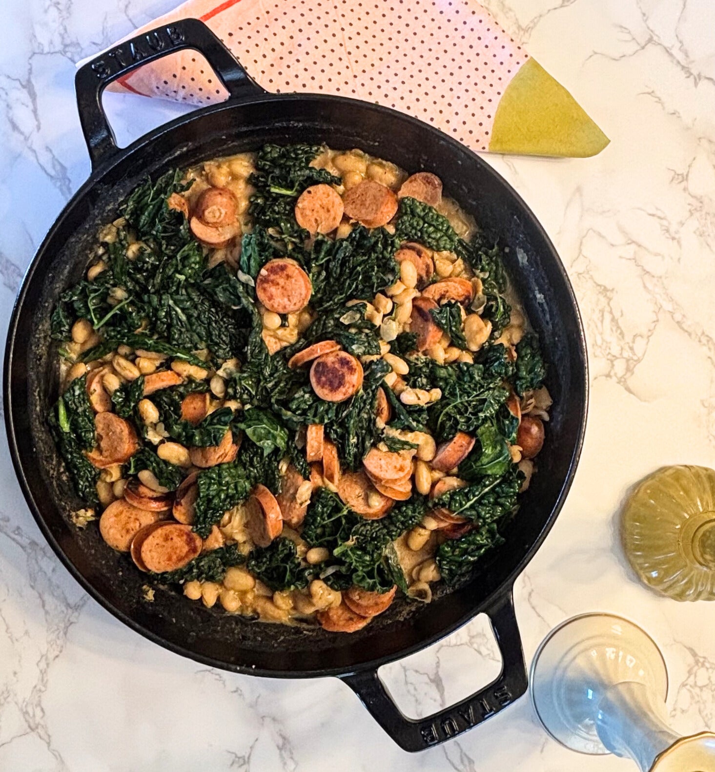 Skillet with greens, sausage, and beans on a marble countertop
