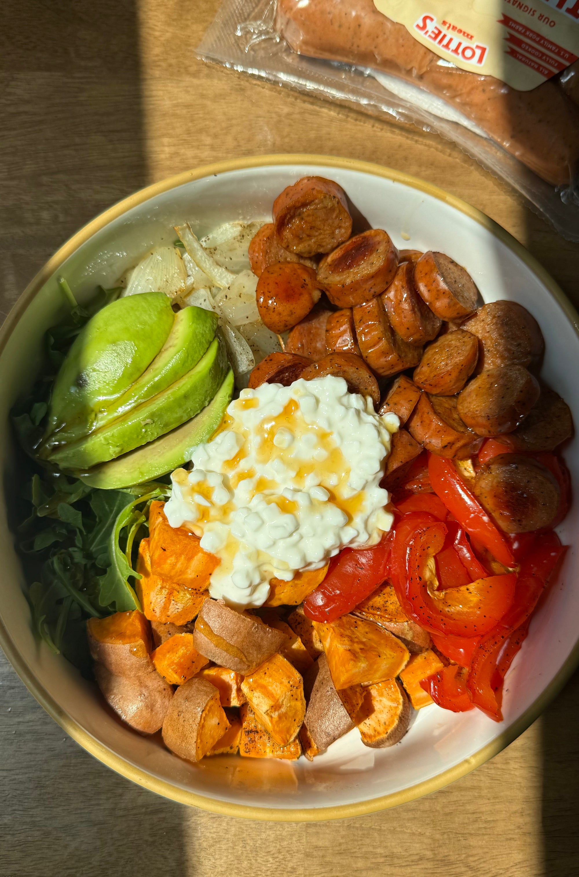 A top-down view of a prepared food bowl containing sliced sausage, sweet potatoes, red peppers, avocado, greens, and a white cream cheese topping with hot honey drizzle, set on a wooden surface with a package of sausage partially visible in the background.