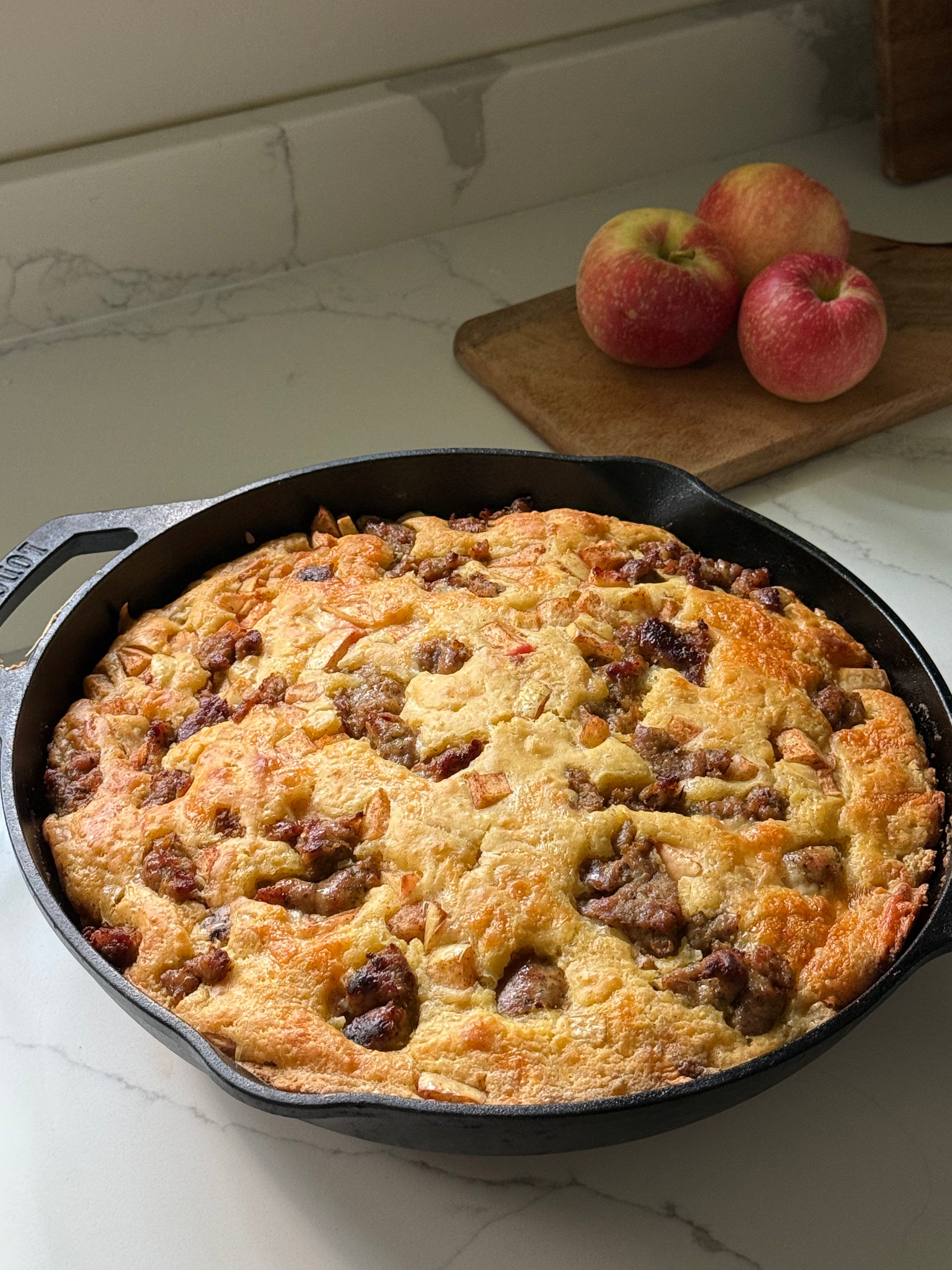 A freshly baked sausage and apple skillet pancake in a black cast iron skillet, set on a white marble countertop with three red apples on a wooden cutting board in the background.