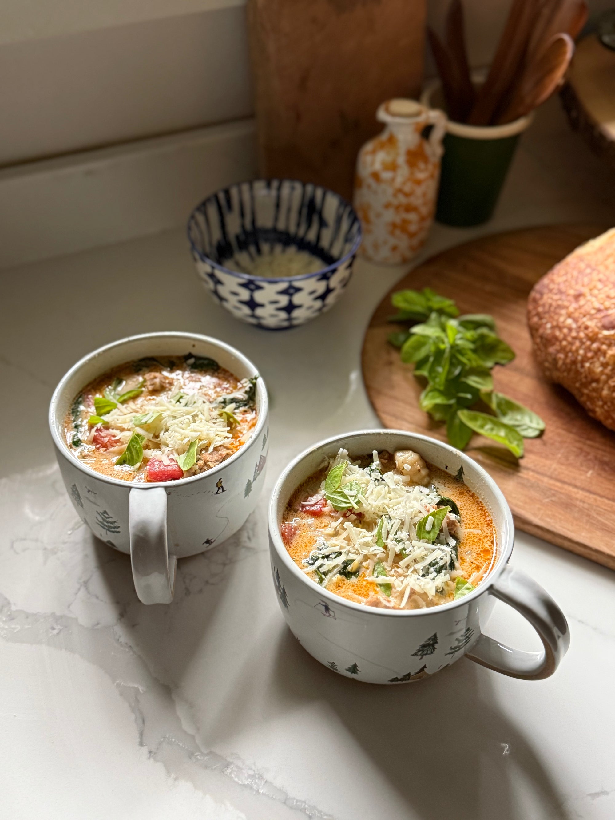 Two white ceramic mugs filled with sausage, gnocchi and spinach soup, garnished with cheese, placed on a white marble countertop in a kitchen setting with bread and basil in the background.