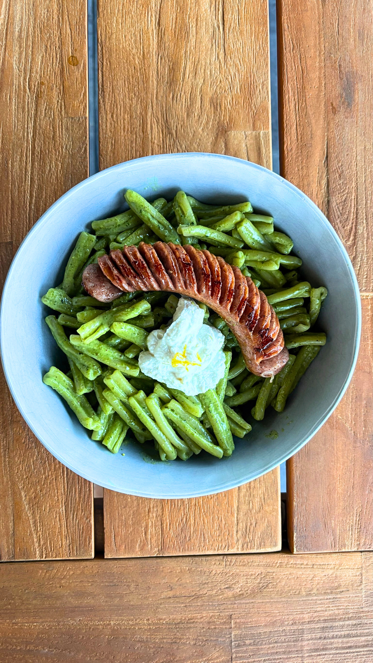 A top-down view of a light blue bowl filled with green pesto pasta noodles, a sliced sausage, and a dollop of whipped ricotta cheese on a wooden table.