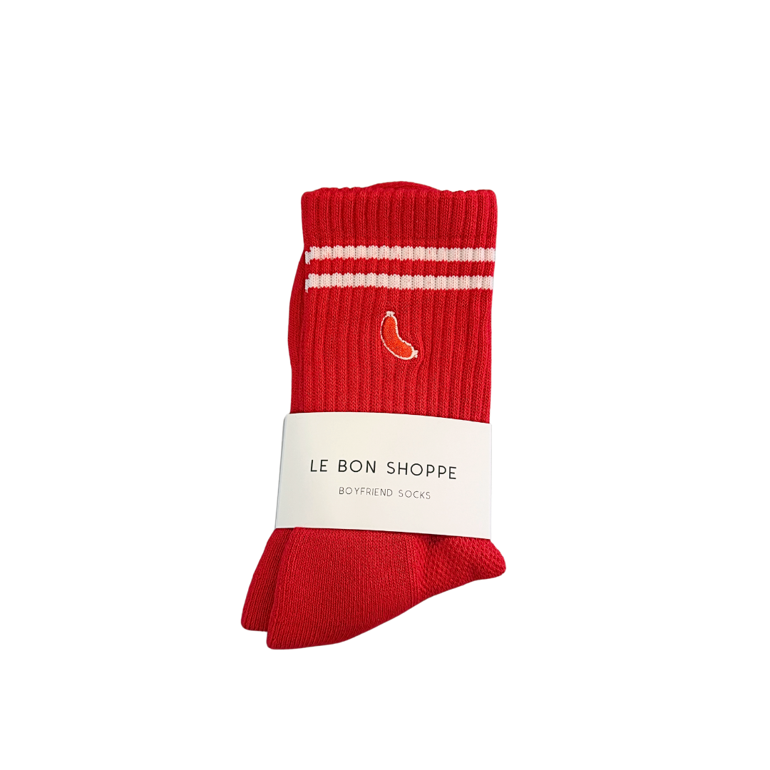 A pair of red crew socks with two white horizontal stripes near the cuff, a small embroidered orange chili pepper logo, and a white paper band reading 'LE BON SHOPPE BOYFRIEND SOCKS', shown on a plain white background.