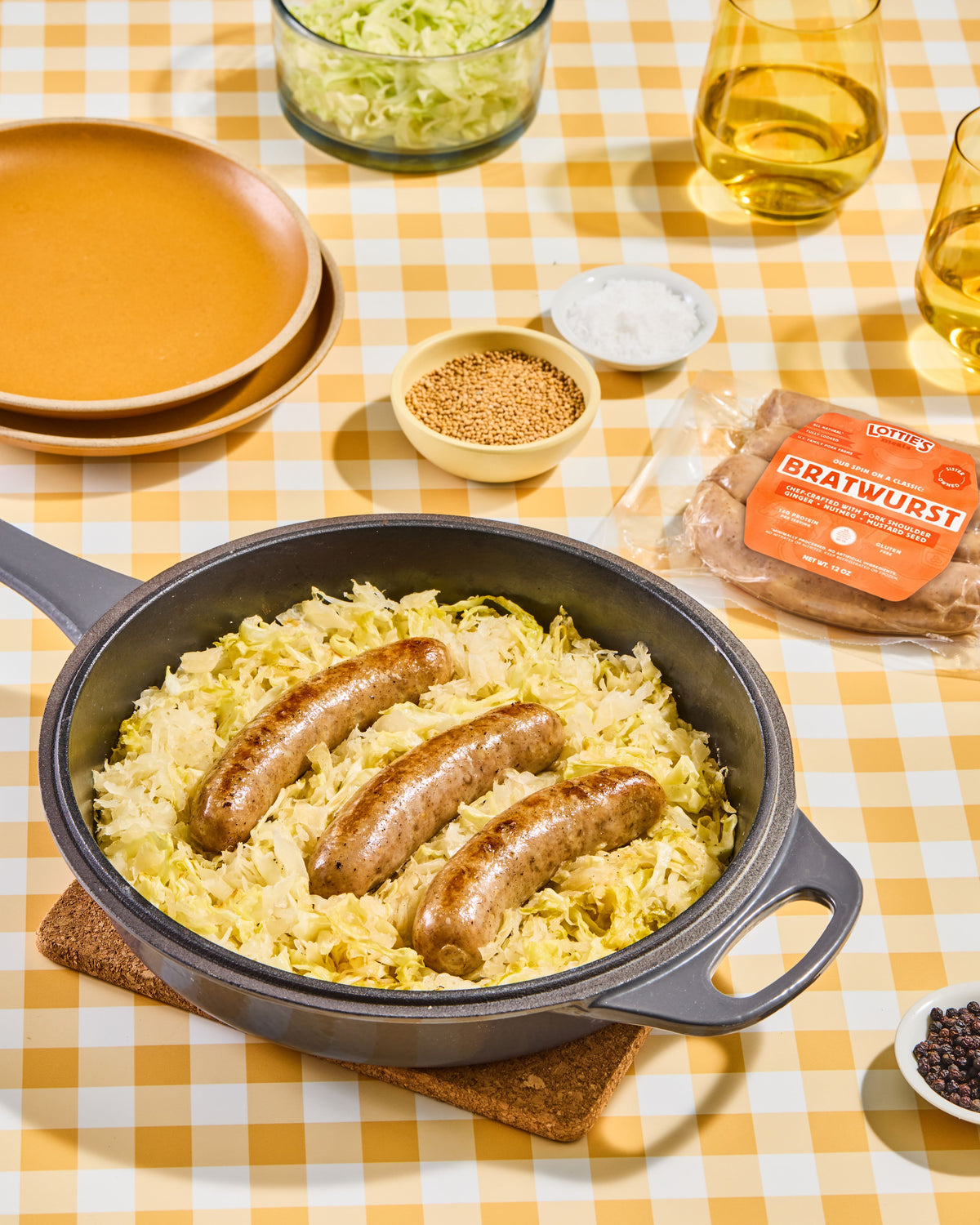 A cast iron skillet filled with braised sausages and sauerkraut on a yellow-and-white checkered tablecloth, surrounded by dining props including plates, glasses, and a package of Brauwurst sausages.