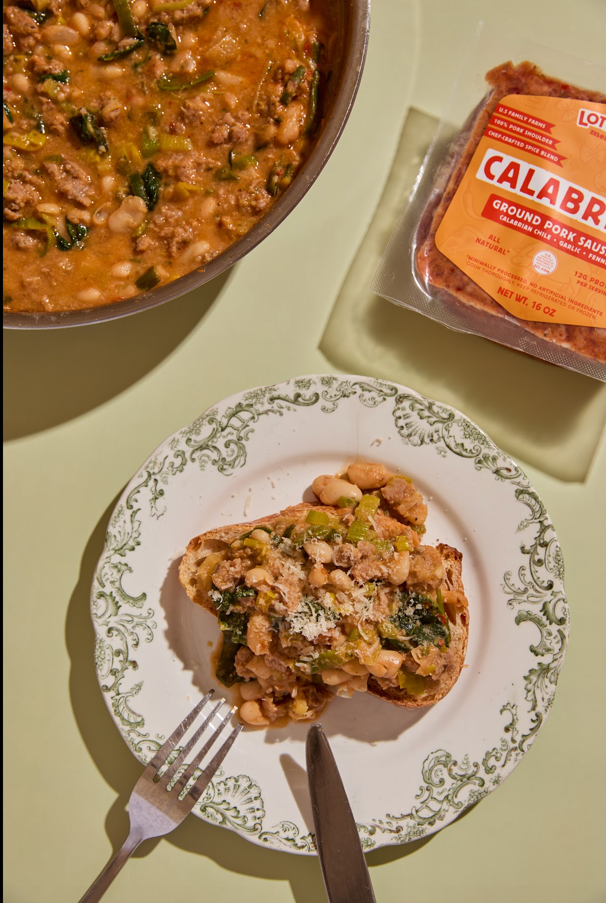 Plate of food with a fork and knife, next to a container of Lot 10 Calabrian ground pork sausage on a light green background.