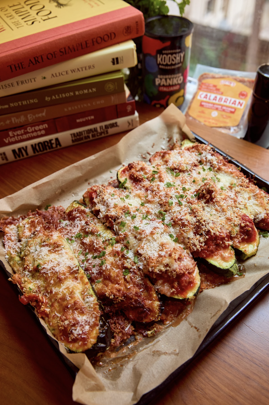 Baked stuffed zucchini boats topped with tomato sauce, melted cheese, and parmesan, served on a baking sheet lined with parchment paper on a wooden table. A stack of cookbooks and a bottle of Kooshy Panko Breadcrumbs are visible in the background.
