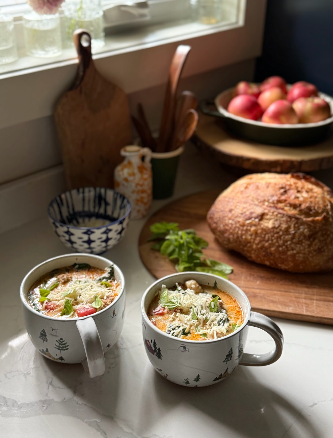 2 white soup mugs on a counter with bread and apples in the background.